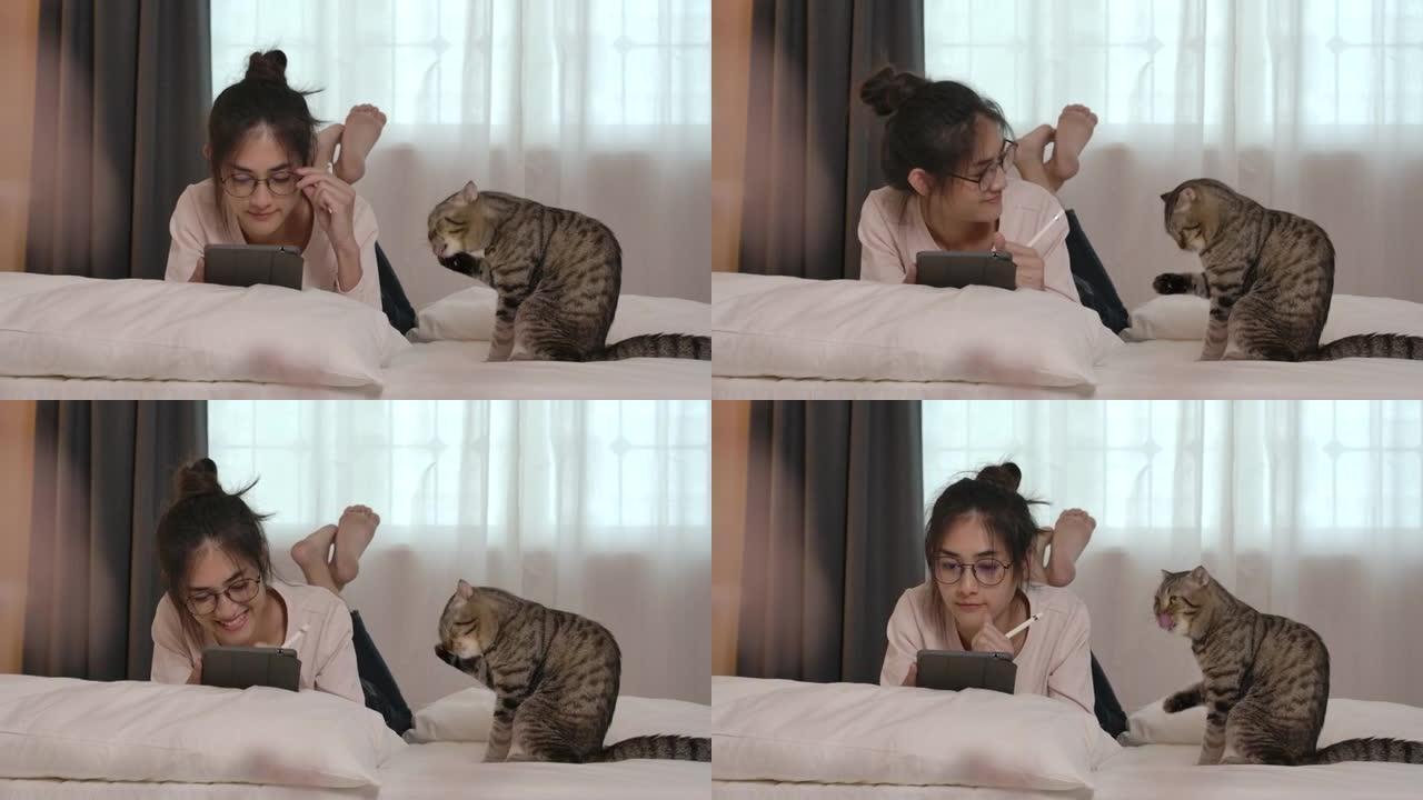 年轻的亚洲眼镜女人躺在床上和可爱的猫。一个漂亮的女孩使用平板电脑在网上工作或学习。检疫期间，穿着休闲