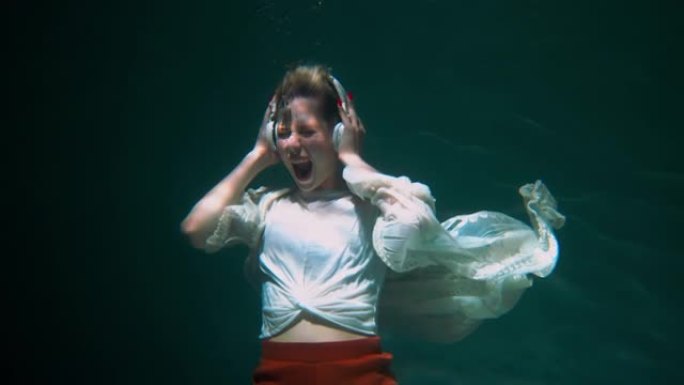 电影在水下拍摄，快乐的年轻美女戴着耳机随着音乐跳舞，穿着波西米亚风的衣服慢动作。