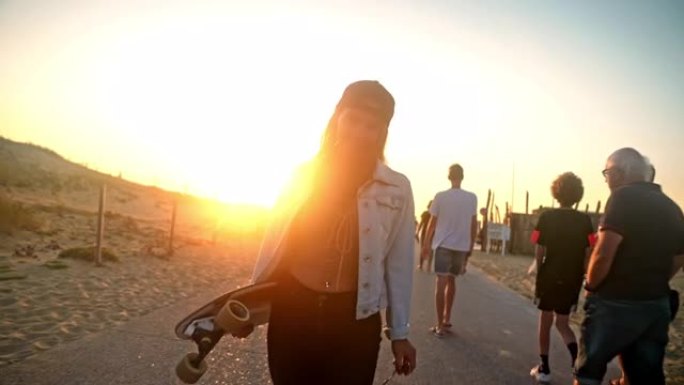 年轻女子，棕色头发，太阳镜在日落时走在木板路上，背着滑板，放下太阳镜