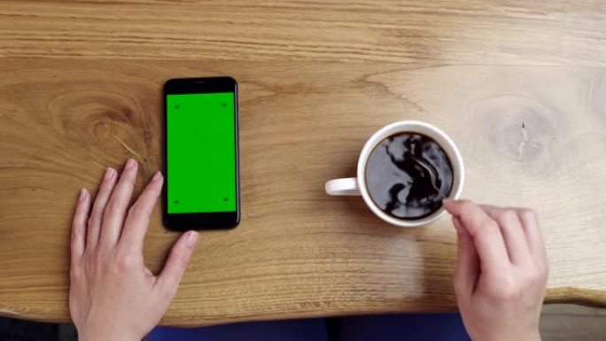 女士无法辨认的女人使用带有色度键绿屏的智能手机，同时用勺子搅拌咖啡