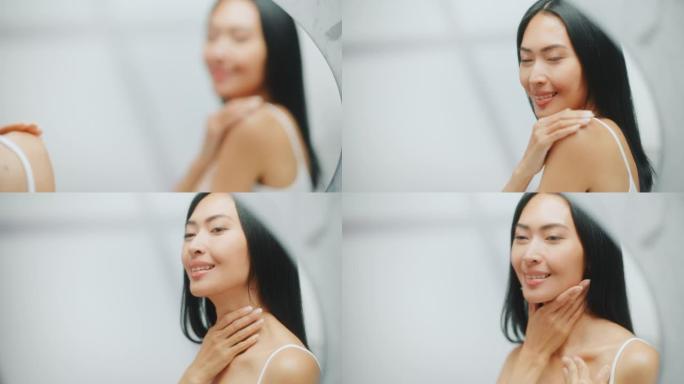 美丽的亚洲女人抚摸着她完美柔软的肩膀，脖子，在镜子里性感地微笑。快乐的女性享受她的美丽。健康天然化妆
