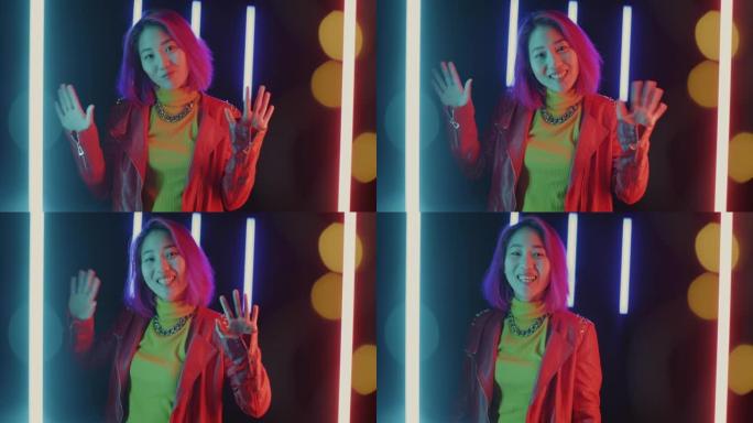 欢乐的亚洲女孩的肖像在霓虹灯照明的黑暗空间中挥舞着双手和微笑的问候