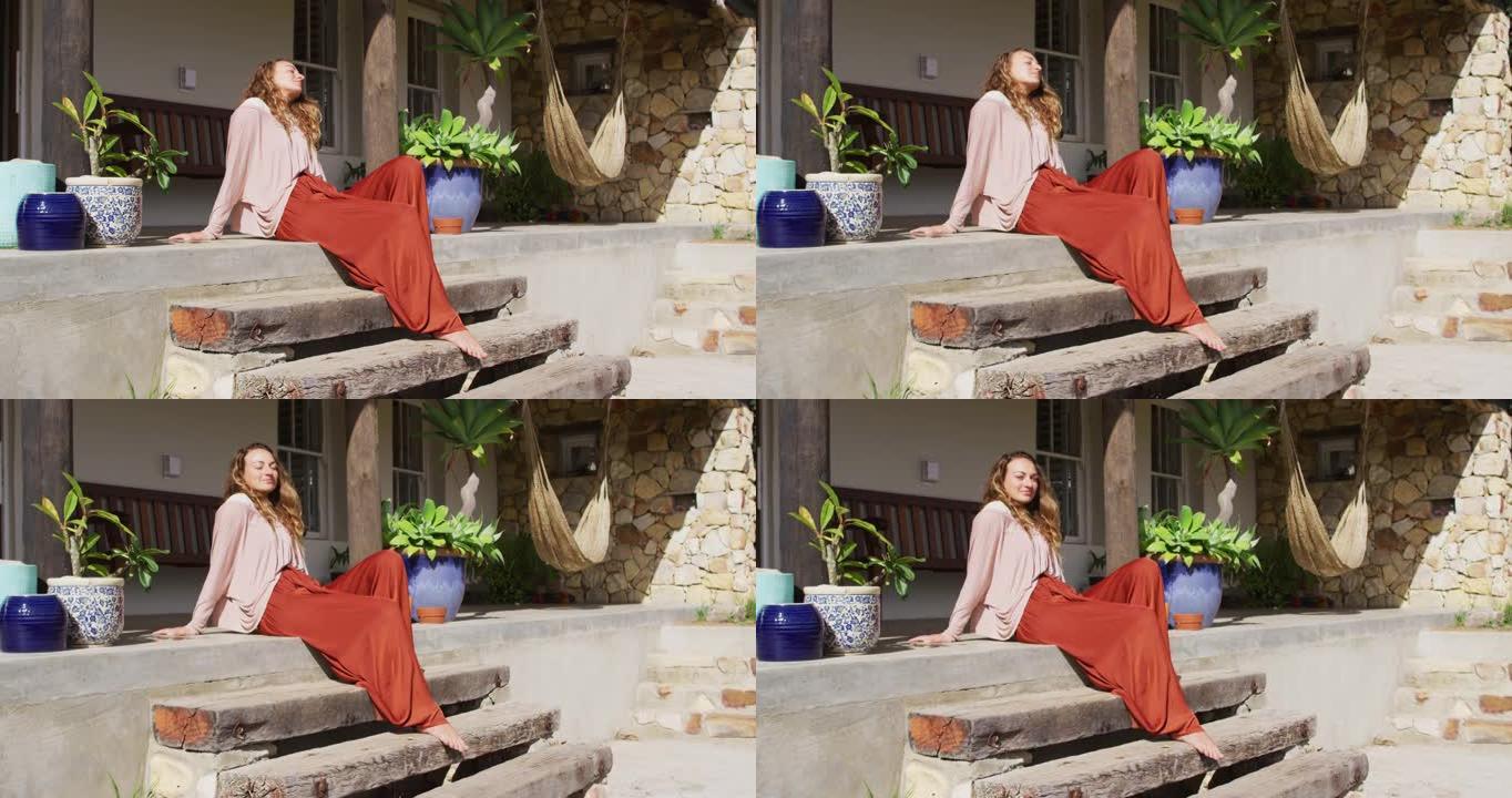 快乐的高加索女人坐在台阶上，在阳光明媚的小屋露台上闭着眼睛放松