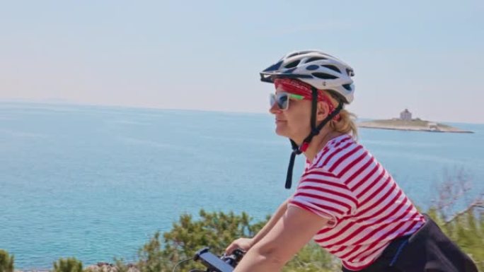 特写镜头金发女子在赫瓦尔岛海岸的一条路上骑自行车，背景是小岛，女子举起手臂