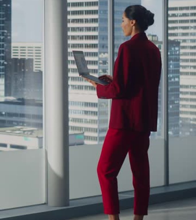 垂直屏幕。成功的女商人站在办公室时使用笔记本电脑，看着大城市的窗外。自信的女性数字企业家开发电子商务