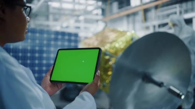 女工程师在进行卫星建设时使用绿屏数字平板电脑。航空航天局: 科学家为太空探索任务组装航天器。超过肩膀