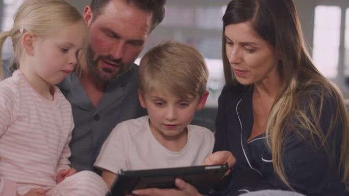 穿着睡衣的微笑家庭坐在沙发上一起使用数字平板电脑玩得开心