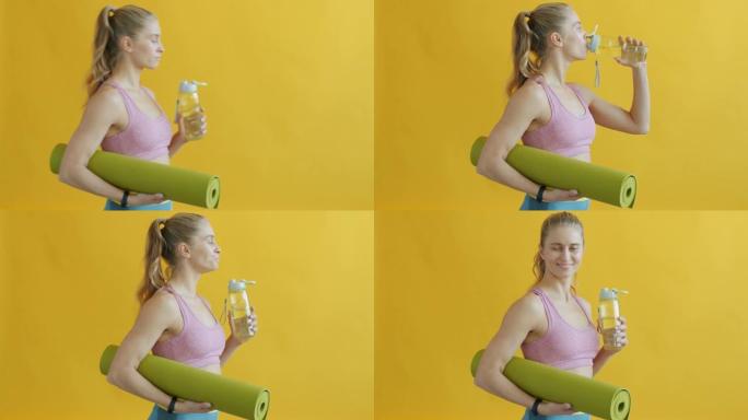 年轻女子用瑜伽垫从可重复使用的瓶子里喝水，眨眨眼看着相机