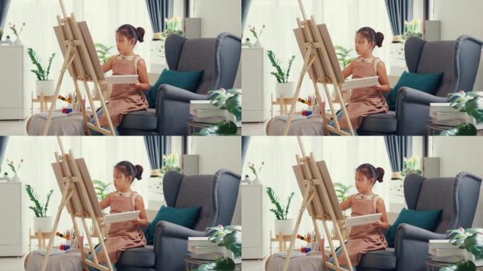 带围裙的亚洲学步女孩坐在沙发椅上，在带调色板的油画画布前使用画笔专注于周末在家画抽象。