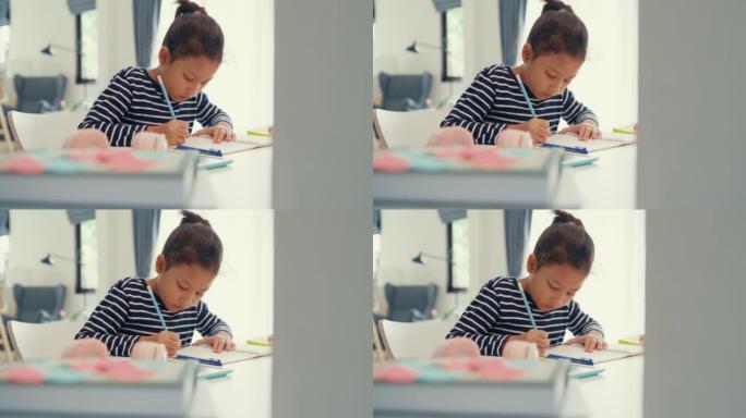 穿着毛衣的亚洲蹒跚学步的女孩坐在办公桌前，拿着记事本，用铅笔专注于写笔记本，周末在家做在线学习课程的