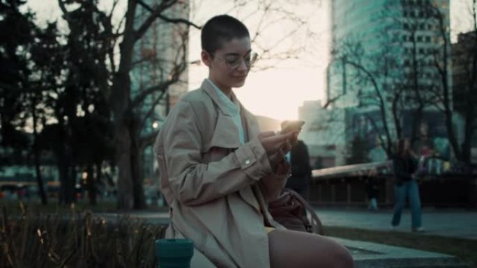 年轻体贴聪明的短发学生妇女使用手机手机应用程序在春天日落公园长凳慢动作。