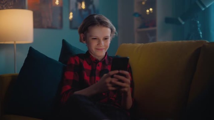 可爱的小男孩坐在黑暗舒适的房间的沙发上，在家使用智能手机。快乐的青少年在线浏览内容，在社交媒体上与朋