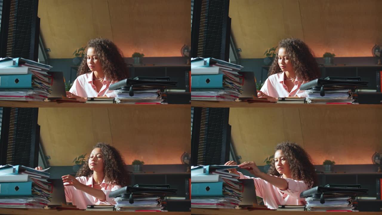 工作中的女首席执行官美丽的混血老板女商人使用笔记本电脑，在装满文件的办公室桌子上检查文件。