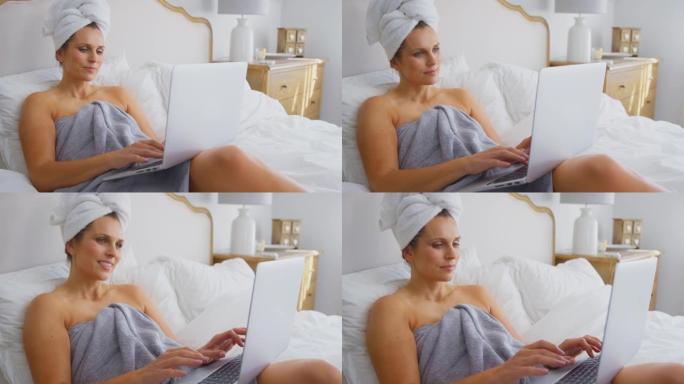 成熟的女人躺在床上，穿着毛巾在家在笔记本电脑上工作