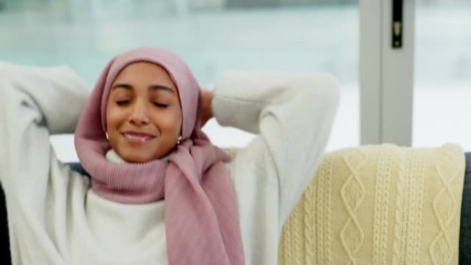 一个穆斯林妇女独自坐在家里客厅的沙发上，放松、自由和快乐。微笑、休息室和生活方式，一位年轻的伊斯兰女