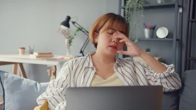 自由亚洲女士坐在沙发上，带着笔记本电脑在线学习，在家里的客厅感到头痛。