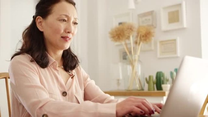 成熟的亚洲女性坐在家里的餐桌旁，用笔记本电脑整理家庭账单和财务 -- 慢动作拍摄