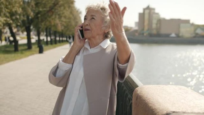 情绪激动的高级女士在手机上聊天并站在城市河边打手势的肖像