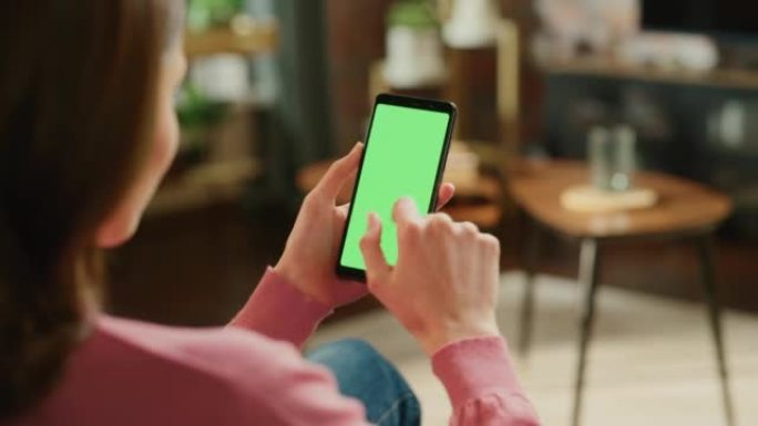 带有绿屏模拟显示的智能手机上的女性手点击显示屏和滚动供稿。女性在家休息，在移动设备上查看社交媒体。特