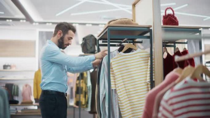 帅气男服装店助理在时尚商场工作。专业商店销售零售助理将具有可持续休闲设计的新彩色系列悬挂在展示架上。