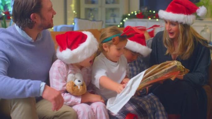 戴着圣诞老人帽子的家庭坐在家里的沙发上打开圣诞礼物