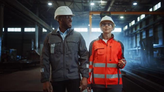 两名戴着安全帽的重工业工程师走进钢铁金属制造厂，进行了讨论。黑人非裔美国工业专家与制造领域的女技术员