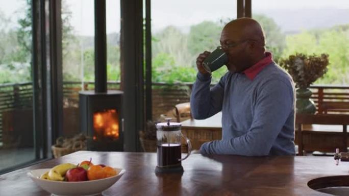 非裔美国高级男子坐在厨房里，可以看到乡村景色，喝一杯咖啡
