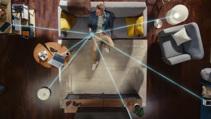 阁楼公寓里的高加索人坐在沙发上，将智能手机连接到方便的智能家居系统的俯视图。VFX动画可视化连接的设