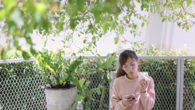 年轻女子坐在大树的树荫下，一边吃零食，一边使用智能手机。