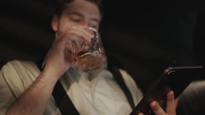一位穿着衬衫和吊带裤的疲惫的经理正躺在沙发上，看着平板电脑上的东西，从波旁威士忌中喝了一口
