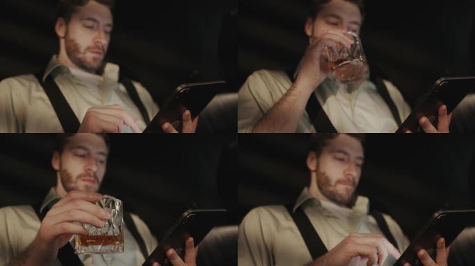 一位穿着衬衫和吊带裤的疲惫的经理正躺在沙发上，看着平板电脑上的东西，从波旁威士忌中喝了一口