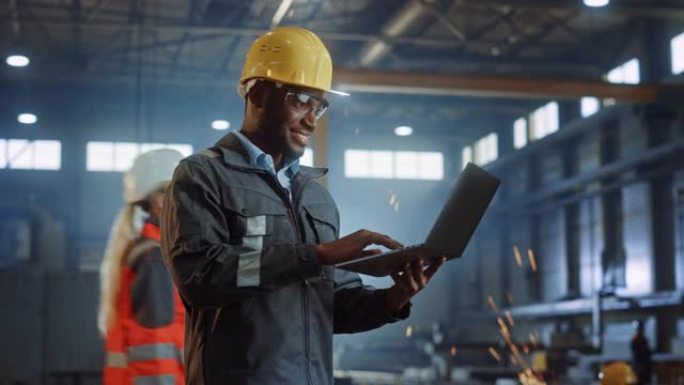 专业重工业工程师/穿着安全制服和安全帽的工人使用笔记本电脑。微笑的非洲裔美国工业专家站在金属建筑制造