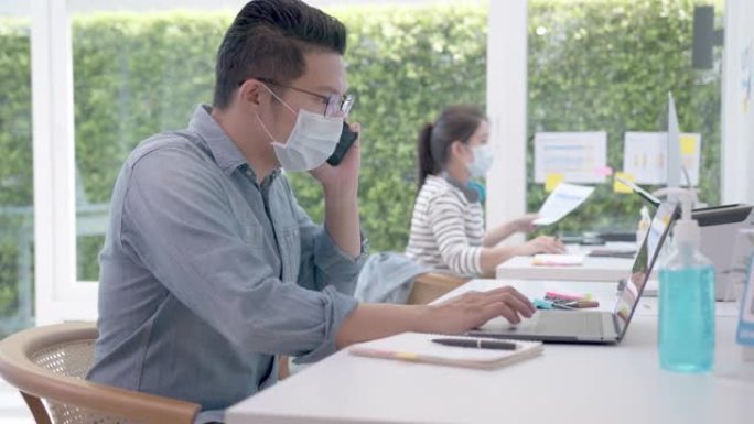 亚洲工人在办公室戴口罩与笔记本电脑一起工作。电晕病毒危机后的社会距离衡量生活方式。在办公室一起工作的