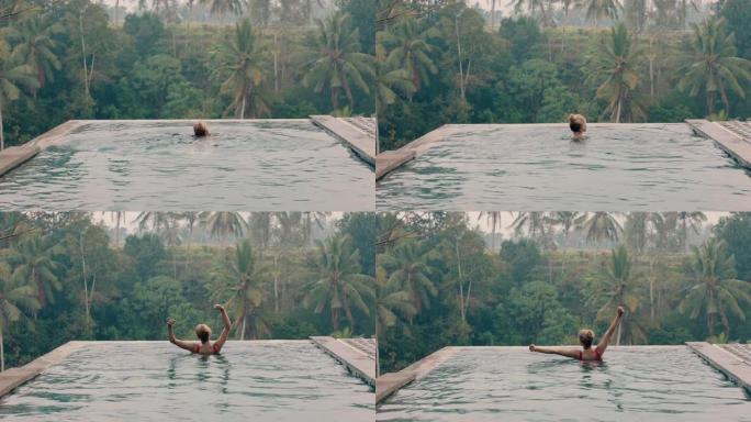 快乐的女人在无边泳池游泳，早上游泳，庆祝在酒店水疗中心的异国度假举起的手臂，可以看到薄雾中的热带丛林