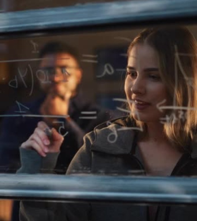 垂直镜头: 白人年轻女子使用可擦除记号笔求解窗玻璃上的数学方程式。白人女导师撰写物理理论并向男生解释