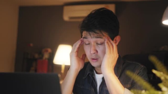 在家庭办公室工作到很晚时，使用笔记本电脑的亚洲男子感到困倦，眼睛上有压力或沮丧