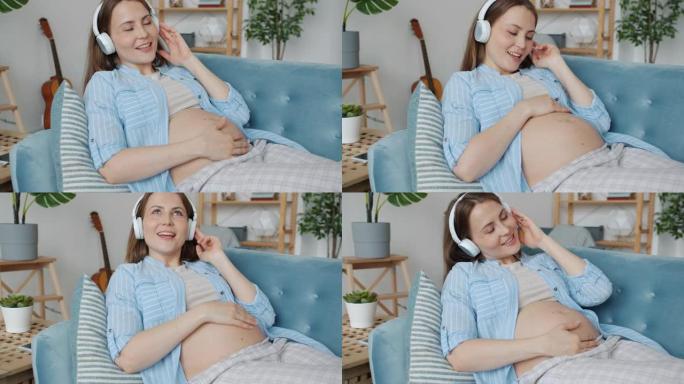 年轻的孕妇戴着耳机享受音乐在家里的沙发上玩得开心