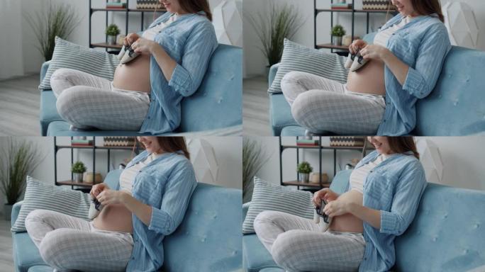 未来的母亲在家里玩婴儿靴和在沙发上微笑的慢动作