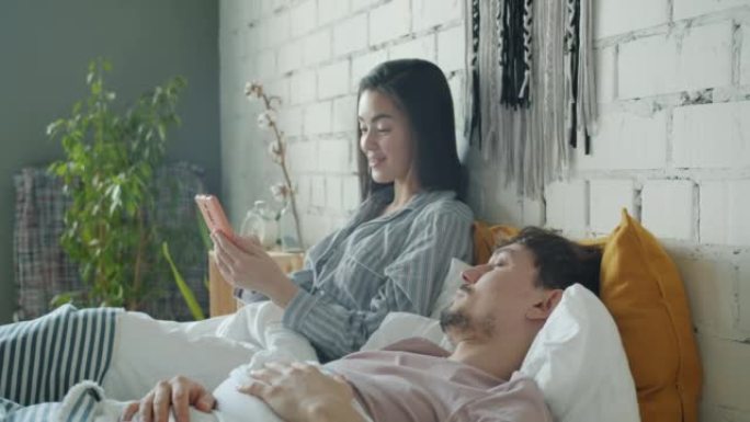 亚洲女子使用智能手机触摸屏微笑，而混血儿男子睡在家里的床上