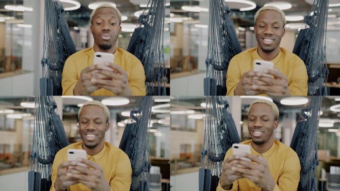 快乐的非洲裔美国员工使用智能手机微笑坐在现代办公室的吊椅上