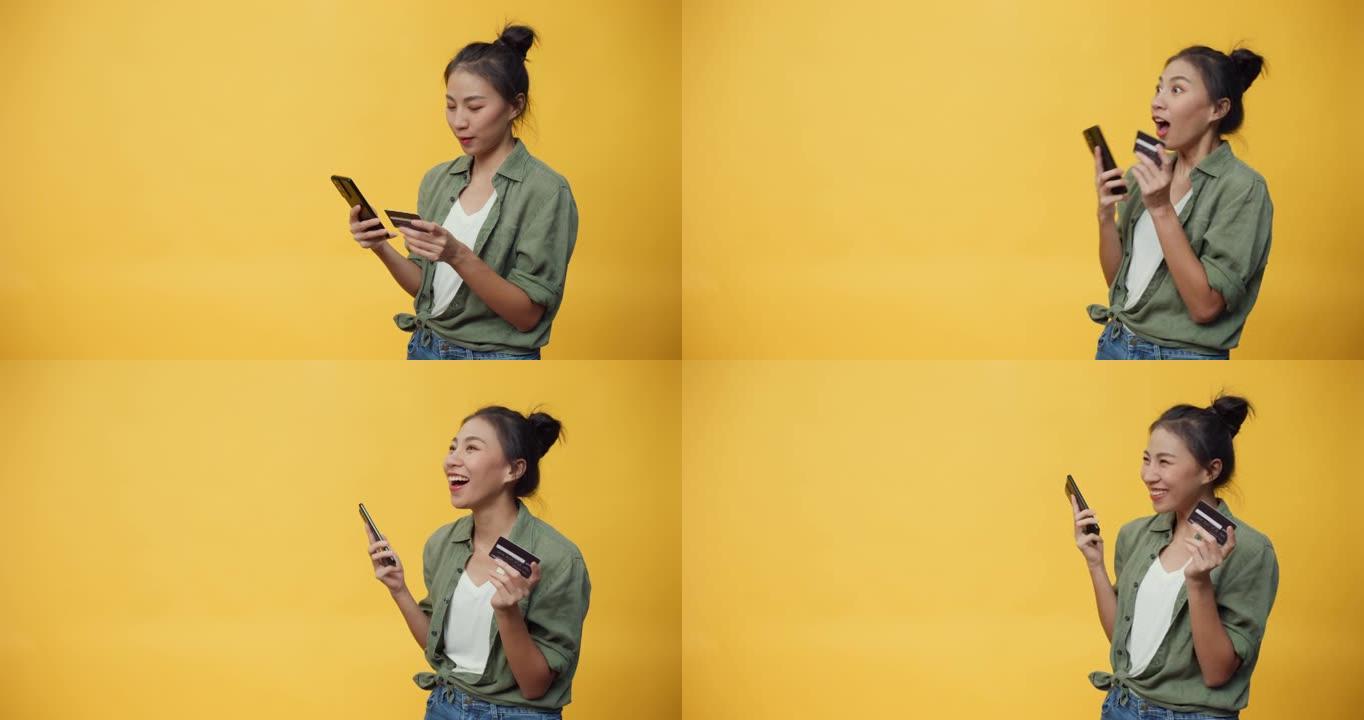 美丽迷人的亚洲青年孤立女士享受手机滚动电子商店和信用卡购买网上商店，成功支付黄色背景。