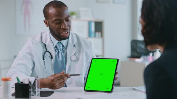 非裔美国医生正在健康诊所用绿屏平板电脑向患者解释诊断。穿着白大褂的助理正在医院办公室阅读病史。