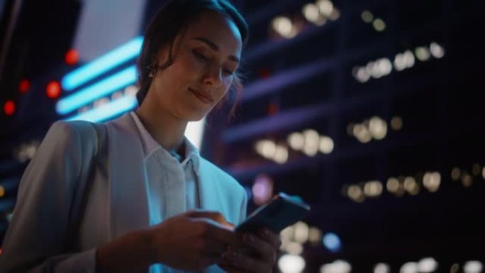 美丽的年轻女子使用智能手机走过充满霓虹灯的夜晚城市街道。使用手机的华丽微笑女性肖像。低角度倾斜向上拍
