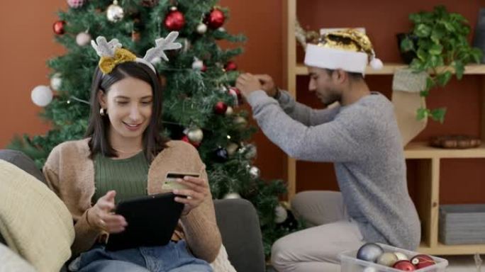 4k视频片段，一名妇女使用数字平板电脑在圣诞节最后一刻购物，而丈夫则在家里装饰树