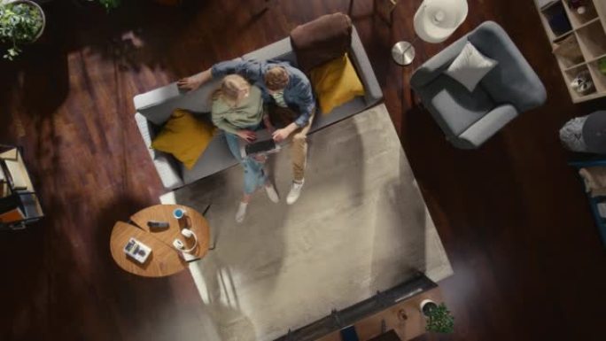 顶景公寓: 恋爱中的幸福夫妇在时尚的客厅使用笔记本电脑，拥抱并抬头看相机。在沙发上: 女友和男友使用