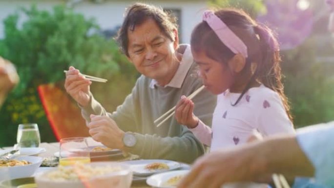 骄傲的祖父在后院吃家庭午餐时看着孙女吃饭。年轻女孩一边享受亚洲美食，一边与爱爷爷在户外学习使用传统筷