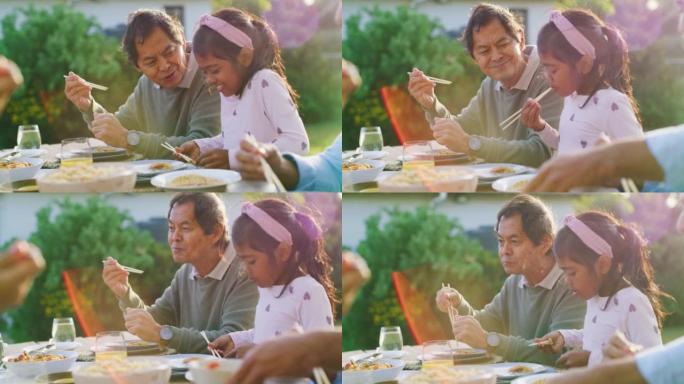 骄傲的祖父在后院吃家庭午餐时看着孙女吃饭。年轻女孩一边享受亚洲美食，一边与爱爷爷在户外学习使用传统筷