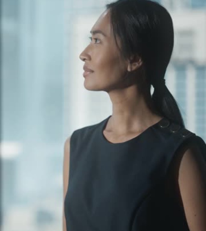垂直屏幕: 穿着黑色连衣裙的女商人在现代办公室里行走，使用智能手机，在摩天大楼的大城市中眺望窗外。自