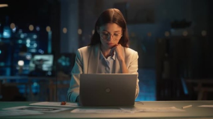 穿着眼镜的时尚女性晚上在公司办公室的笔记本电脑上工作。年轻的经理浏览互联网，在线购物并阅读朋友和同事