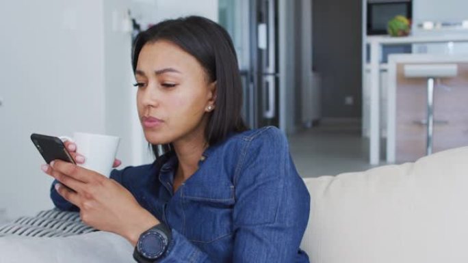 混合种族妇女坐在沙发上使用智能手机喝杯咖啡